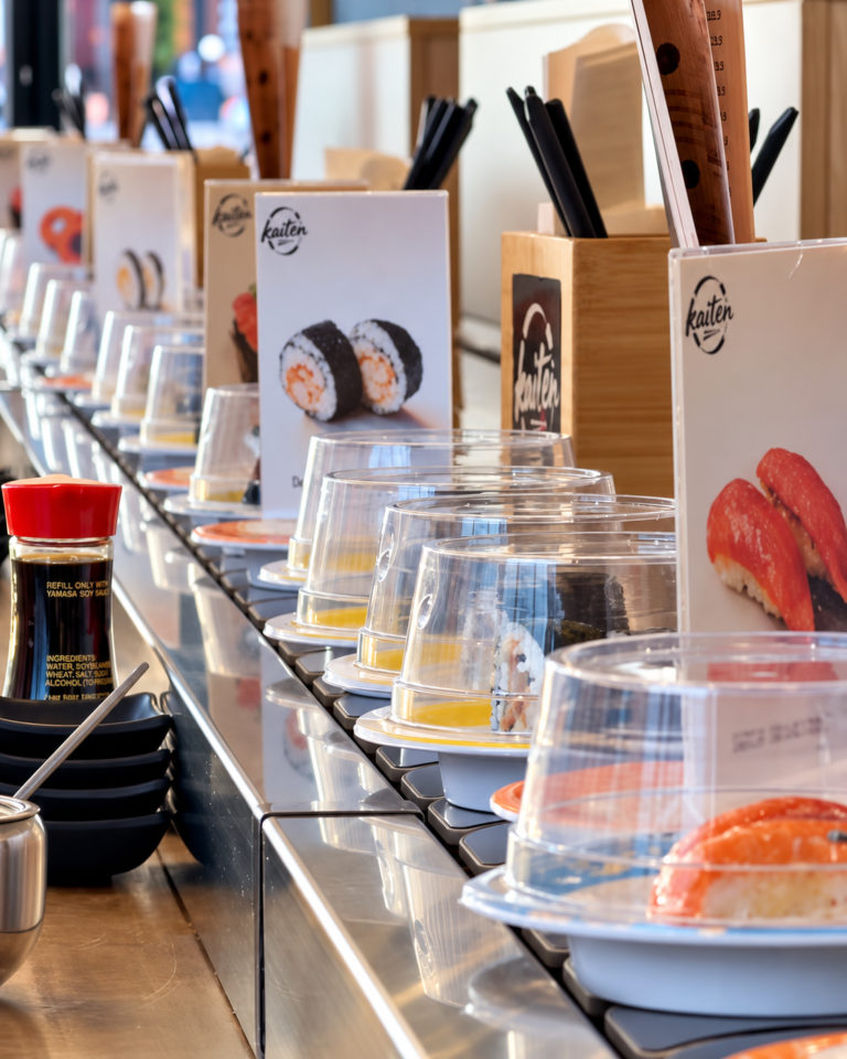 Covered sushi plates passing menu cards and chopstick holders on Kaiten's sushi train